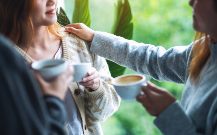 Three people are standing in a circle with coffee cups in their hands. One person has placed their hand on another person’s shoulder.