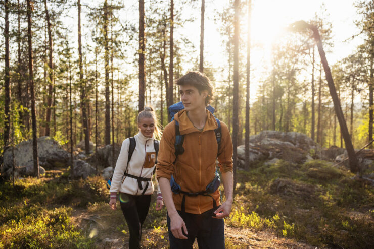 Nuori mies ja nainen kävelevät peräkkäin polulla. Ympärillä näkyy mäntymetsää ja kivikkoa. Henkilöillä on reput selässä. Henkilöiden takaa paistaa aurinko.