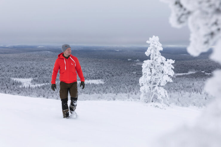 A man wearing a red hoodie from Lapland University of Applied Sciences is walking in the fells. Behind him, a snowy landscape unfolds. In the foreground, trees covered in heavy snow can be seen.