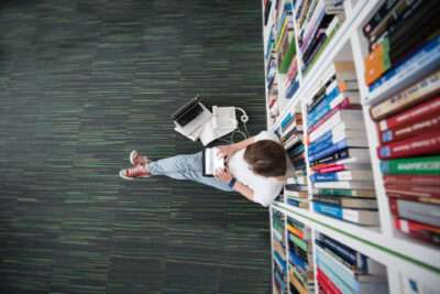 A person is sitting on the floor. Next to them are a laptop, books, and headphones. The person is leaning against a bookshelf. The picture is taken from above.