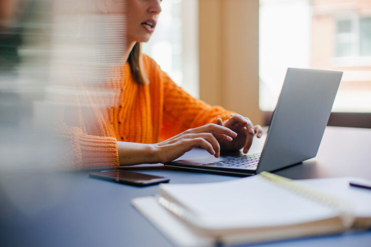 “A woman is working at a laptop. Her fingers are on the keyboard. She is wearing an orange sweater.