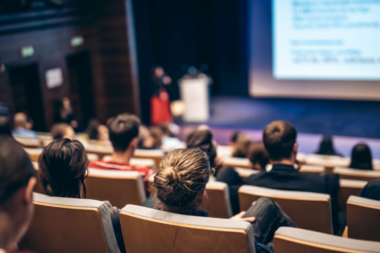 Henkilöitä istuu auditoriossa ja kuuntelevat puhujaa. Henkilöt istuvat selin kameraan. Esiintyjä ja valkokangas näkyy epäterävänä taustalla.