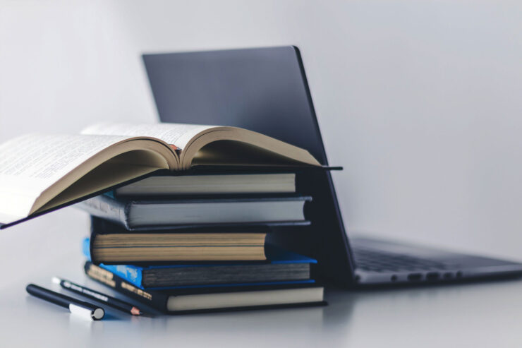 A stack of books and a laptop. The top book in the stack is open. There are pens next to the stack of books.