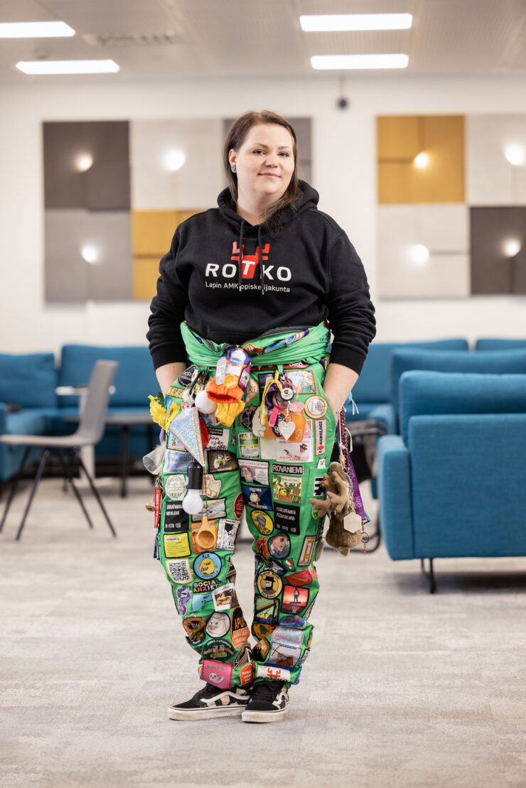 Tummahiuksinen nainen katsoo kameraan hymyille. Hän on kampuksella. Hänellä on yllään opiskelijahaalarit, joihin on ommeltu haalarimerkkejä. Haalareista roikkuu myös pieni poro-pehmolelu.
