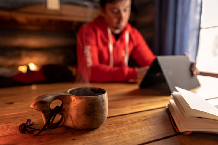 Lapin ammattikorkeakoulun punaiseen huppariin pukeutunut henkilö istuu pöydän ääressä. Hänen edessään on tabletti. Etualalla näkyy kuksa, jossa on höyryävää juomaa.