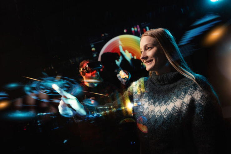 A blonde-haired woman wearing a sweater is pointing at a bright light. In the background, a man with VR glasses on and controllers in his hands can be seen. They are in a dark room with bright, multicolored lights.