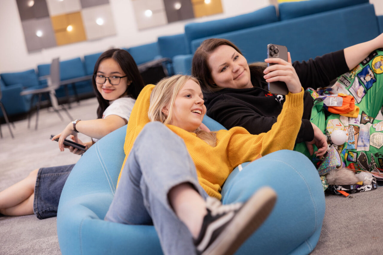 Three students are sitting on campus, relaxed, back-to-back, and looking at a phone screen. Everyone is smiling.