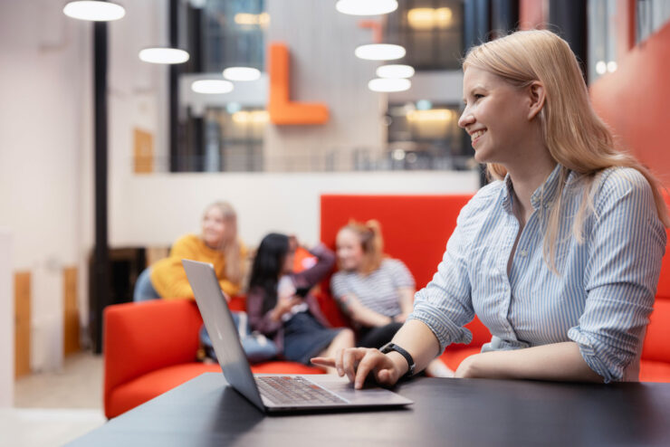 A blonde-haired woman is sitting at a table in the foreground of the picture. In front of her is a laptop. She is looking past the camera and smiling. One hand is on the laptop’s keyboard. In the background, three people can be seen having a conversation out of focus.