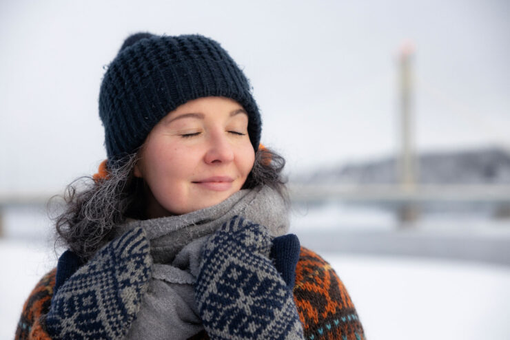 The woman smiles peacefully. Her eyes are closed. She is wearing a hat, mittens, and a sweater. In the background, a snowy landscape is visible, blurred.