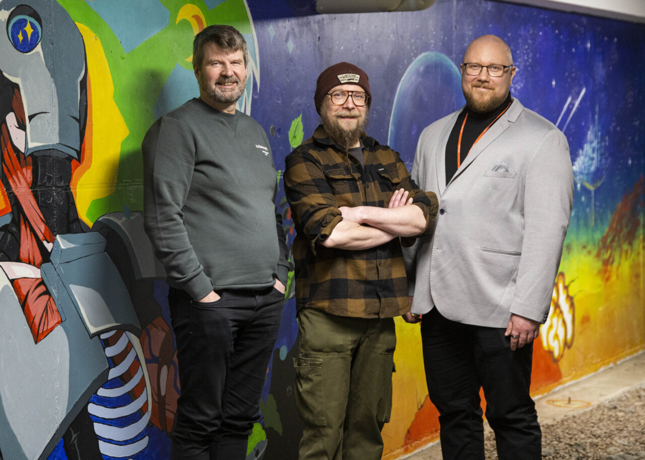 Kolme henkilöä poseeraa värikkään graffitiseinän edessä. Seinän maalauksessa näkyy suuri robotin hahmo sekä avaruus- ja luontoteemaisia elementtejä. Henkilöt seisovat vierekkäin käytävässä, jonka lattia on sorapintainen, ja taustalla oleva taideseinä luo elävän ja visuaalisesti vahvan tunnelman.