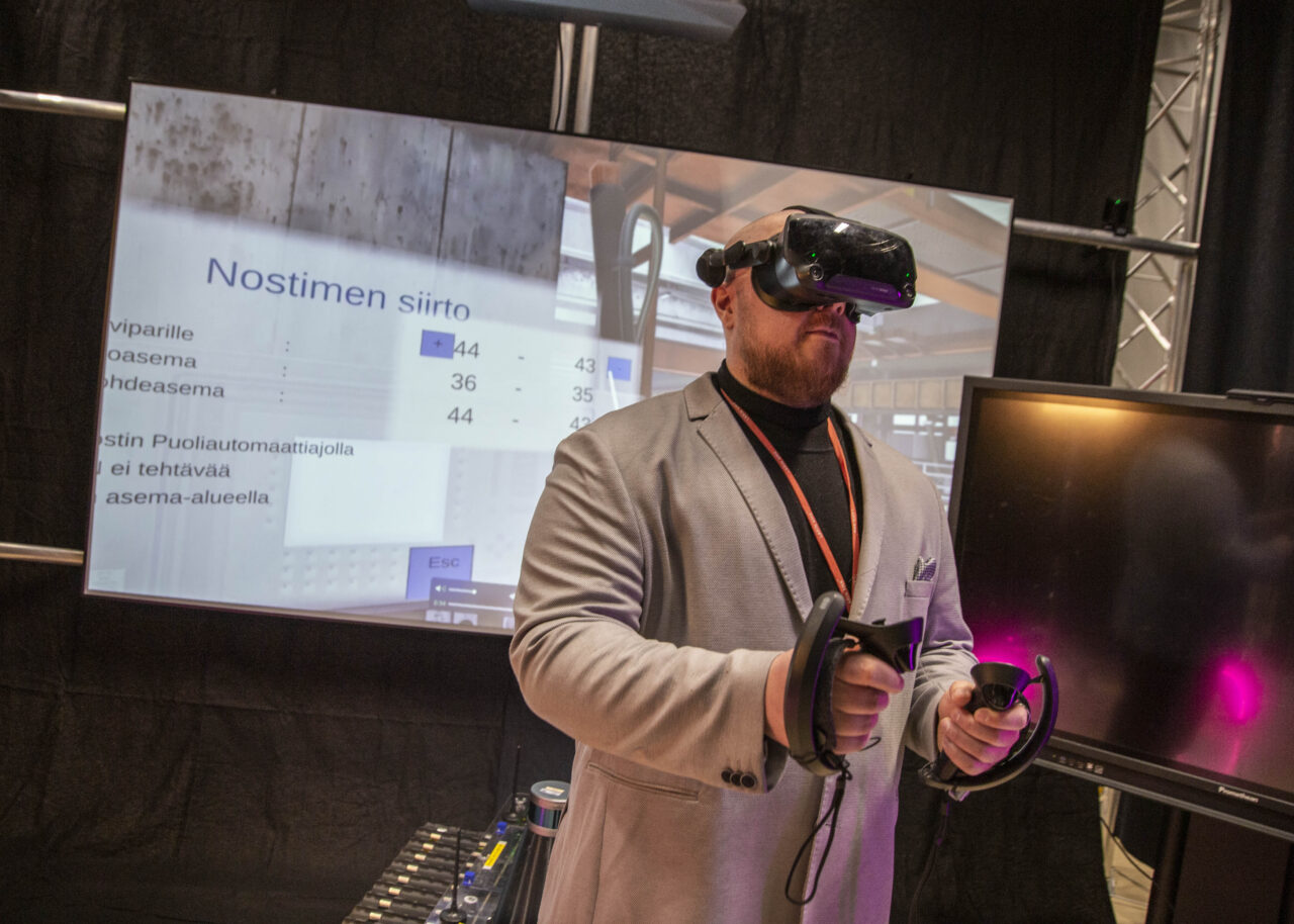 Henkilö käyttää VR-laseja ja käsiohjaimia teknologiaesittelyssä. Taustalla näkyy suuri näyttö, jossa on käyttöliittymä ja tekstiä, muun muassa otsikko “Nostimen siirto”. Näyttö esittää ohjauspaneelin kaltaista näkymää. Henkilö seisoo valaistussa messu- tai demotilassa, jossa on myös muita näyttöjä ja laitteistoa.