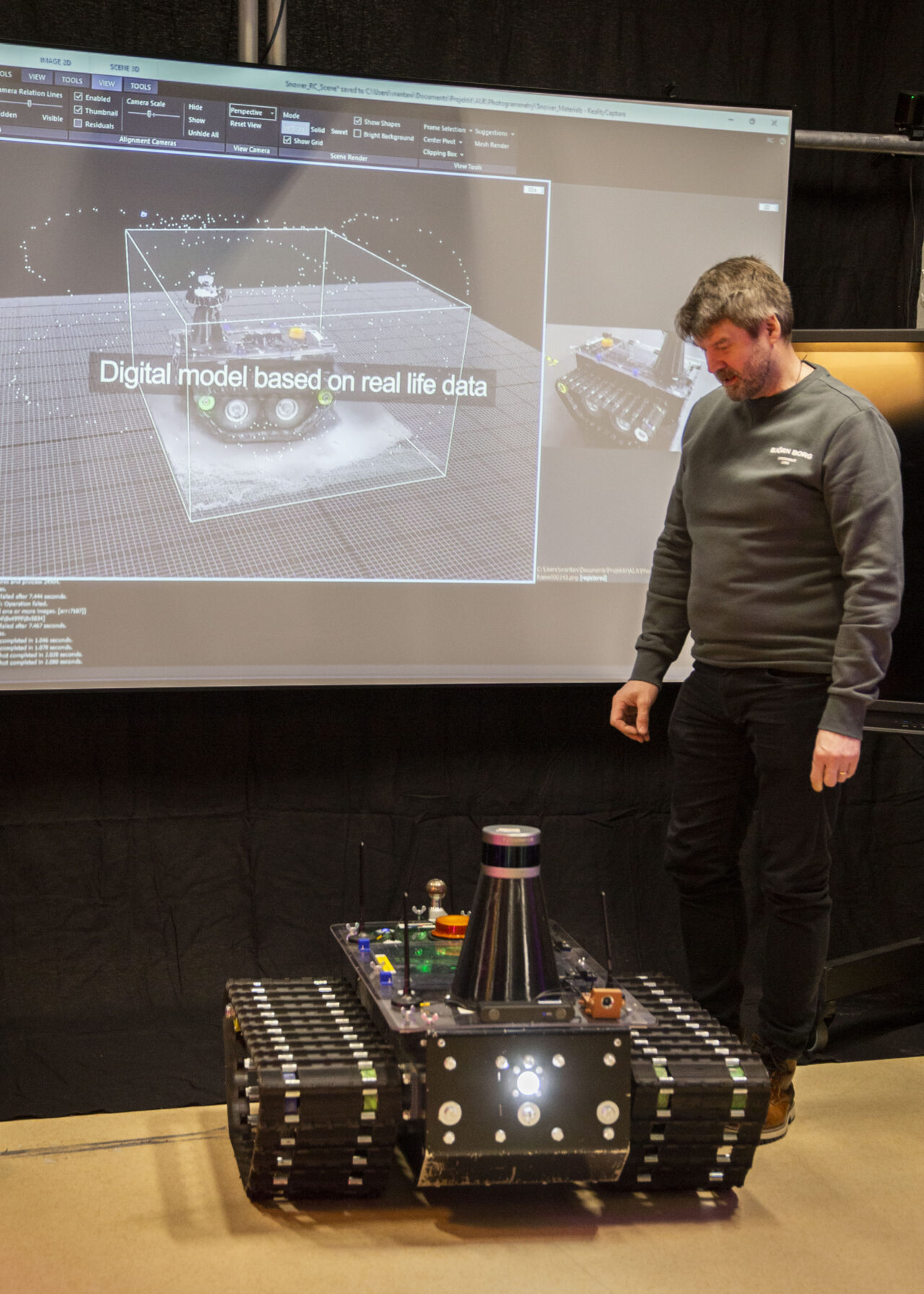 Henkilö seisoo teknologiaesittelyssä tela-alustaisen robottilaitteen vieressä. Taustalla on suuri näyttö, jossa näkyy digitaalinen 3D-malli samasta laitteesta, ja ruudulla lukee “Digital model based on real life data”. Robotissa on erilaisia antureita, valoja ja komponentteja, ja näyttöympäristö luo vaikutelman demonstraatiosta tai messutilanteesta.