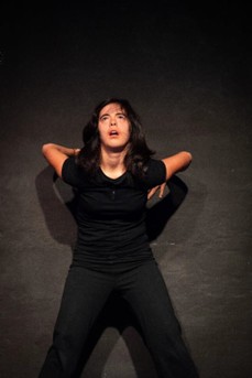 Angela, a young woman with Down Syndrome shown while dancing. She has long dark hair. She is wearing black clothes. She is looking up and not at the camera. Angela has her arms behind her body, resting against the wall and pushing her forward.