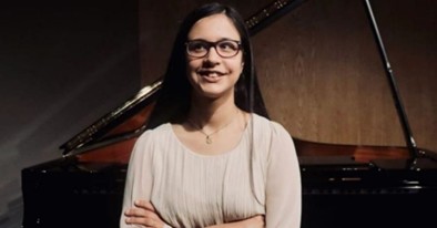 Alessia, a young girl with autism is standing in front of a black piano. She is wearing glasses and has long dark hair. Alessia has her arms crossed over her chest. She is wearing a light pink top and is smiling at the camera.