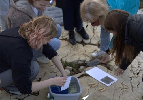 Figure 11. A photograph shows several people gathered outdoors on cracked ground, leaning over a container filled with liquid. One person dips a paper into the liquid while others observe, and nearby papers and small sticks are placed on the ground in this hands-on activity.