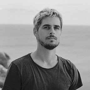 Niels, a young man. He has short hair. He is looking straight at the camera. Behind him there is the sea. He is wearing a dark t-shirt.