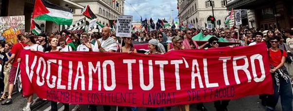 Figure 18. A photograph shows a large crowd of people marching in a city street between tall buildings. At the front, participants hold a long red banner with large white text reading “VOGLIAMO TUTT’ALTRO,” while others behind carry flags and signs, forming a dense demonstration scene.