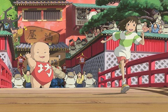 An animated scene shows a child running across a red bridge in a busy, colorful town filled with small human-like figures. In the foreground, a large baby-like character wearing a red outfit waves, while in the background a lively crowd gathers around traditional-style buildings with decorative details.