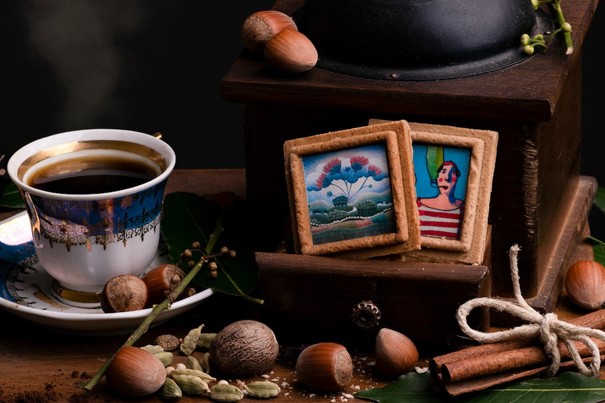 A  still life composition on a wooden surface shows a cup of coffee on a saucer, a vintage grinder, and several nuts and spices scattered around. In front of the grinder are two small artworks embedded into traditional Croatian spicy pepper cookies, alongside cinnamon sticks and green leaves.