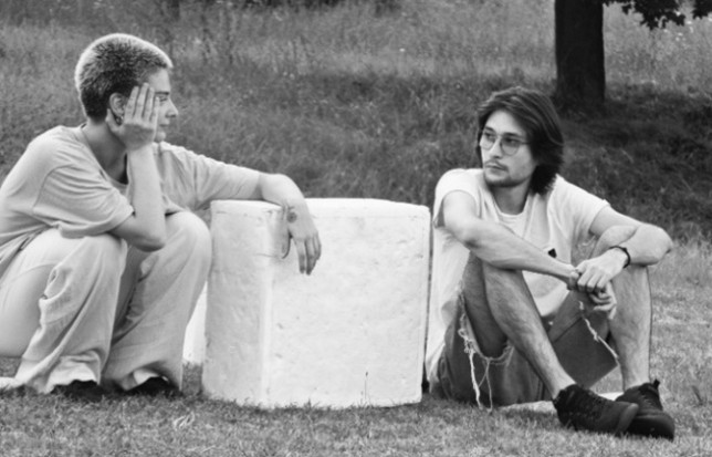 Figure 4. A black-and-white photograph shows two people sitting on grass outdoors. Both are facing each other, one resting a head on a hand and the other sitting with arms around bent knees, with trees and open space in the background.