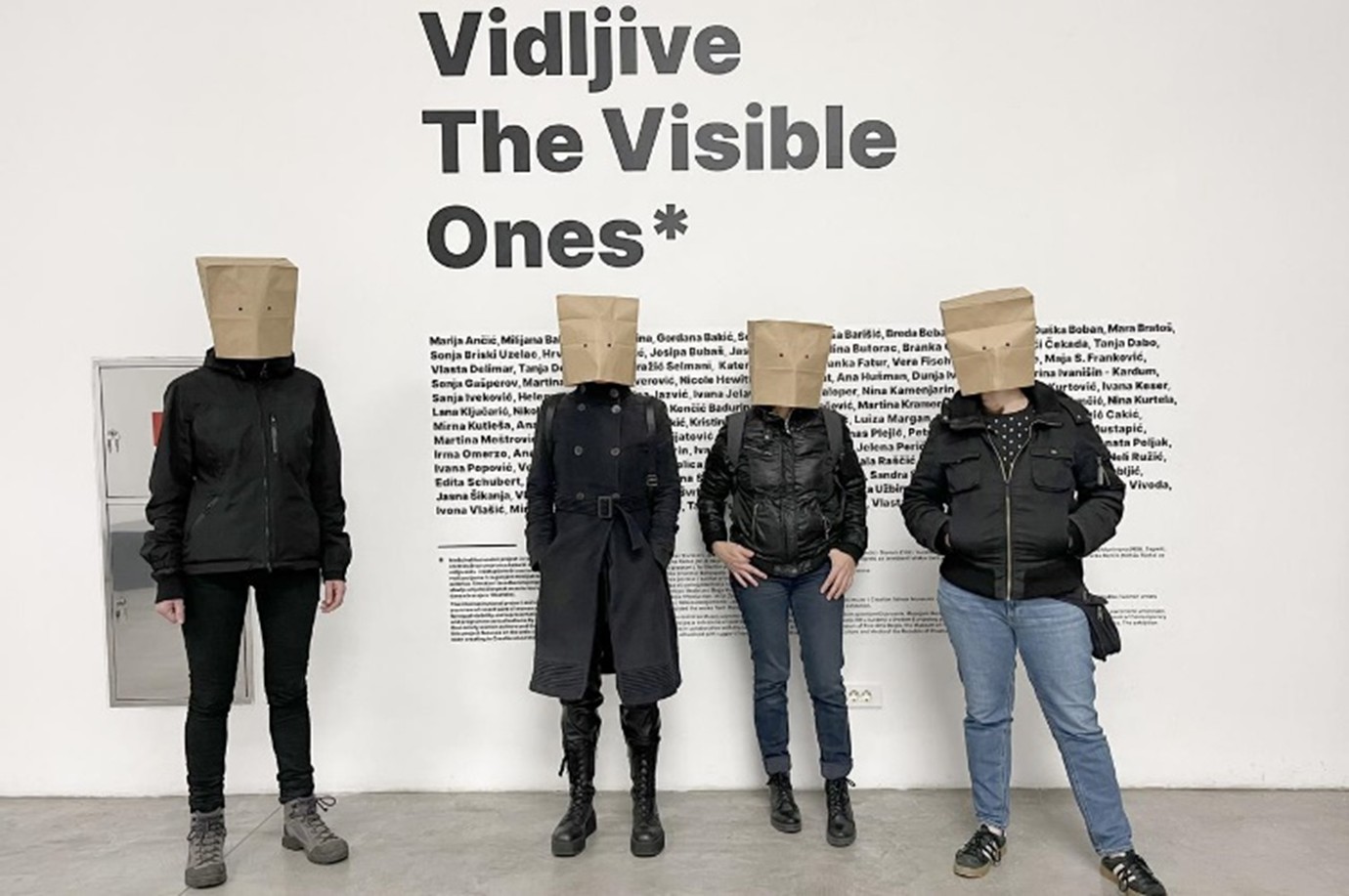 A photograph shows four people standing in a row indoors against a white wall, each with a paper bag covering the head. Behind them, large text on the wall reads “Vidljive - The Visible Ones,” followed by a block of smaller names or text.