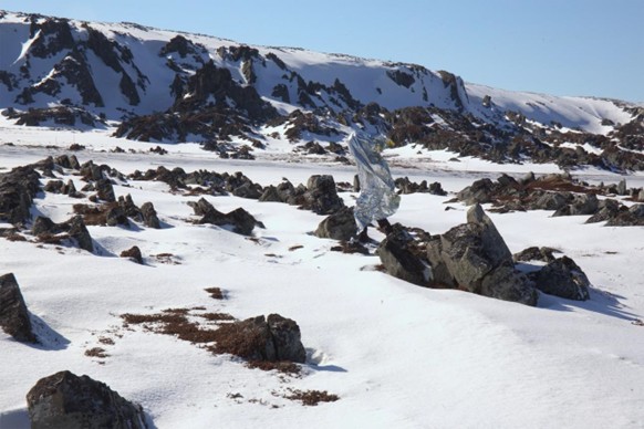 A photograph shows a snowy mountainous landscape with scattered rocks and patches of exposed ground. In the middle of the scene, a person wearing a reflective, foil-like cloak moves among the rocks, standing out against the natural surroundings under a clear blue sky.