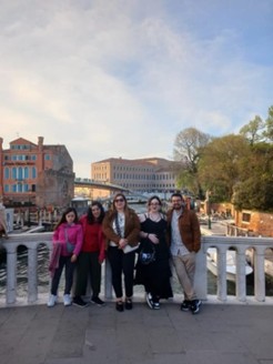 A group of five friends standing together in a line on a bridge with the background of Venice. They are all wearing different colours. They are all smiling straight at the camera. It is late afternoon.