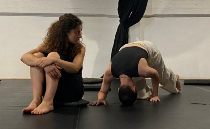 Figure 5. A photograph shows two people in a studio space with a dark floor and curtain in the background. One person sits on the floor with knees drawn up, while the other balances in an inverted pose with hands and head on the floor, forming a curved body shape.
