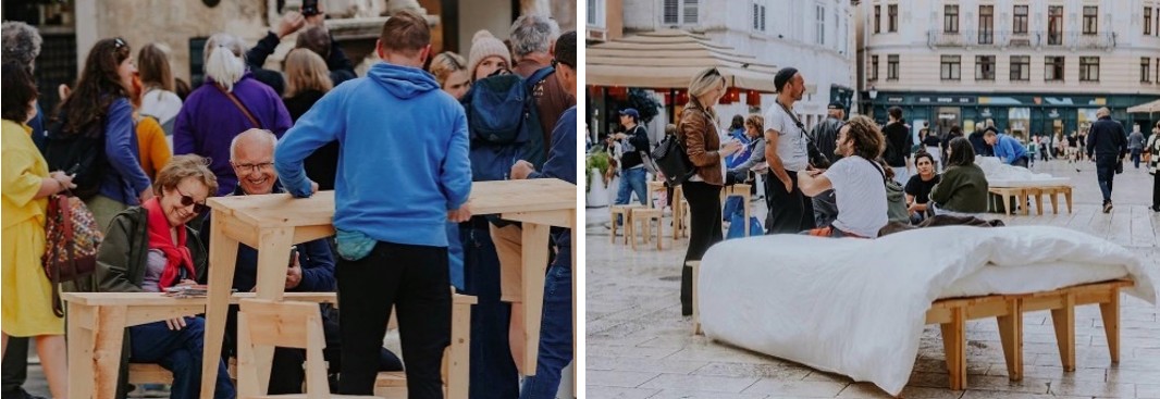 Figure 6. A photograph shows a group of people gathered around tall wooden tables outdoors. In the foreground, two seated individuals interact with materials on the table, while others stand nearby in a busy, social setting with more people and buildings visible in the background. Figure 7. A photograph shows people gathered in a public square with light-colored buildings in the background. In the foreground, individuals sit and stand around simple wooden tables and benches, including one covered with white fabric, while others move through the open space behind them. 
