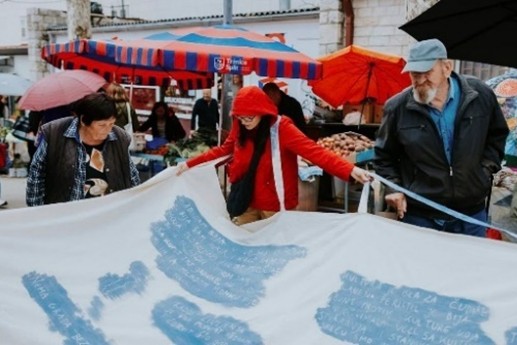 Figure 8. A photograph shows people gathered at an outdoor market with colorful stalls and umbrellas in the background. In the foreground, three individuals hold and unfold a large white fabric printed with blue text, while others stand nearby among market goods and displays.