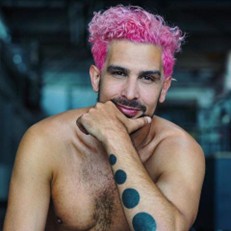 Marcelo has pink hair and smiling into the camera. Marcelo holds his chin with one hand, with blue tattoos of filled circles which can be seen on his arm. On the left- hand side of the image, you can see the Professional Media Presence Project logo which is pink and green