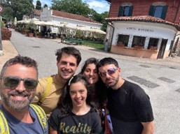 A photograph shows five people standing close together outdoors, taking a selfie. Behind them is an open square with café tables, umbrellas, and buildings with light-colored facades, creating a casual public setting.